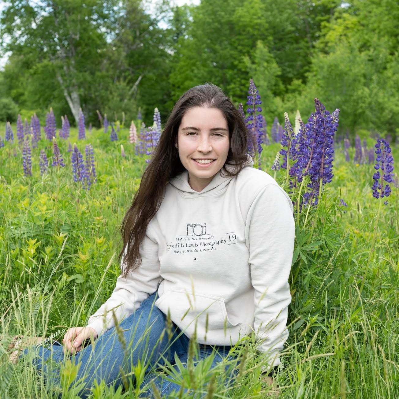 Meredith Lewis sitting in a New Hampshire lupine field, wearing her Meredith Lewis Photography hoodie